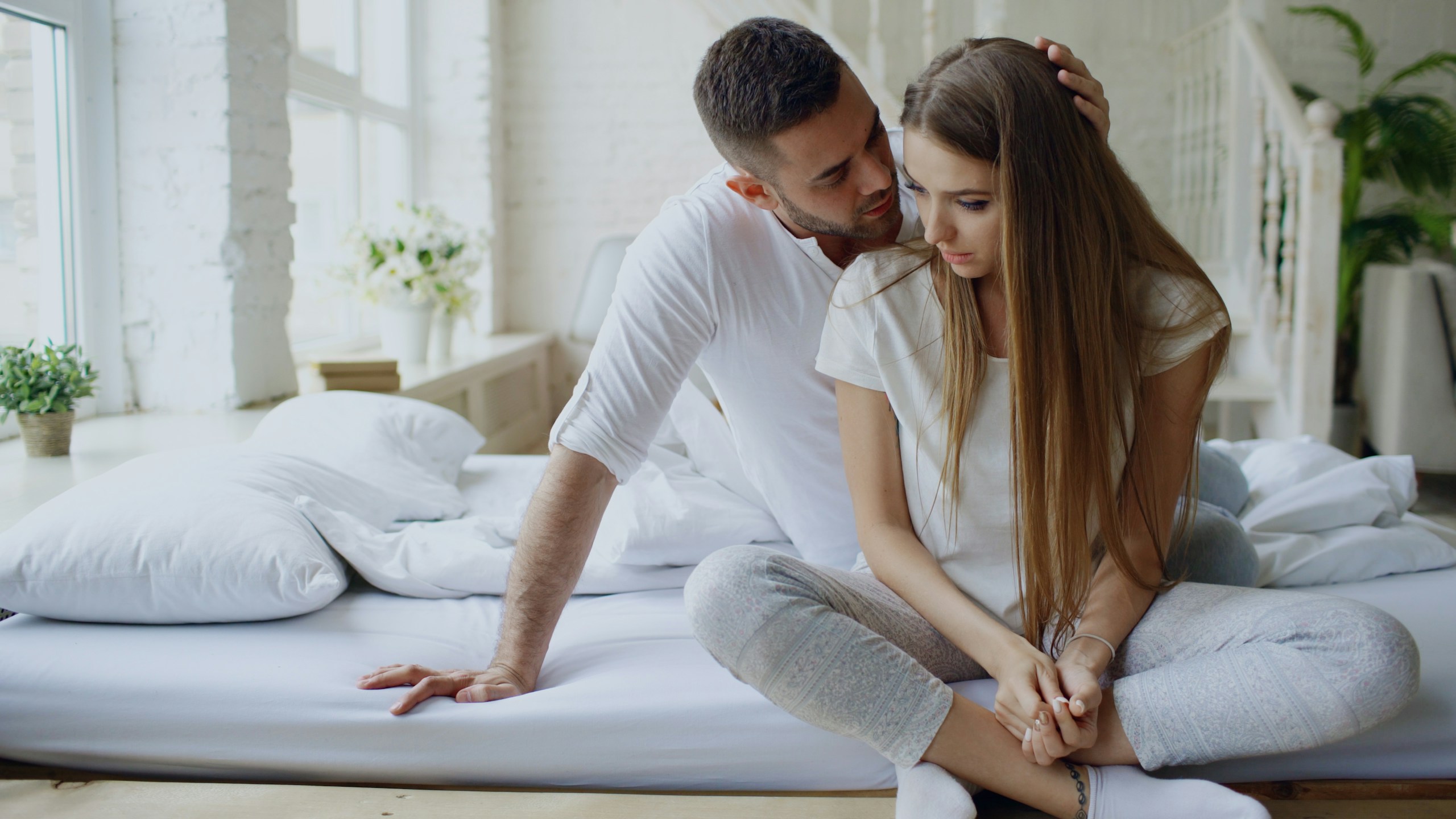 Depressed young woman sitting in bed and crying while her boylfriend come and embrace her in bedroom at home